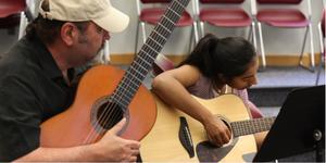 Man in a classroom teachcing a young woman to play guitar
