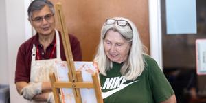 An instructor looking on as a student paints on canvas on her easel.